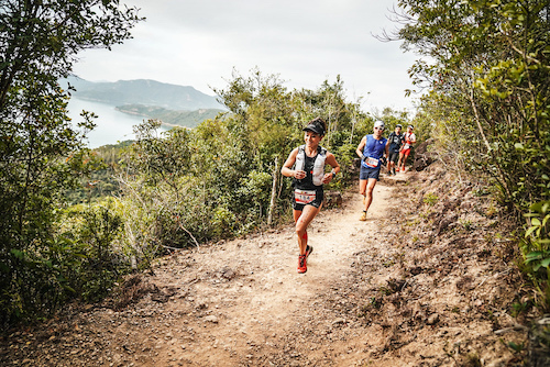 The World Trail Majors Hong Kong 100. A line of people are running along a dusty trail with a lake in the distance behind them