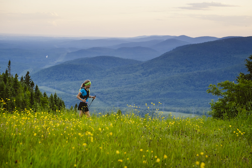 A woman running across a rich green field with wild flowers with forested hills beyond her