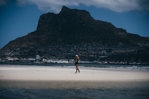 A woman running across a sandy beach with a large lumpy mountain behind her, towering above a city
