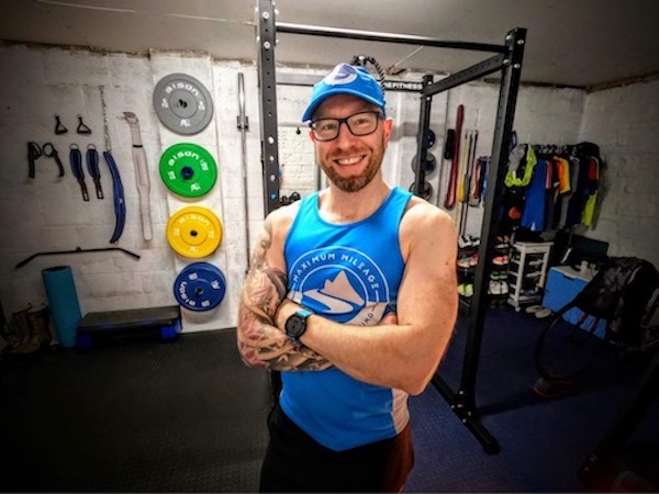  Delving into the world of strength training for ultra runners - a man wearing glasses and a baseball cap standing in a home gym, smiling at the camera, where a blue vest