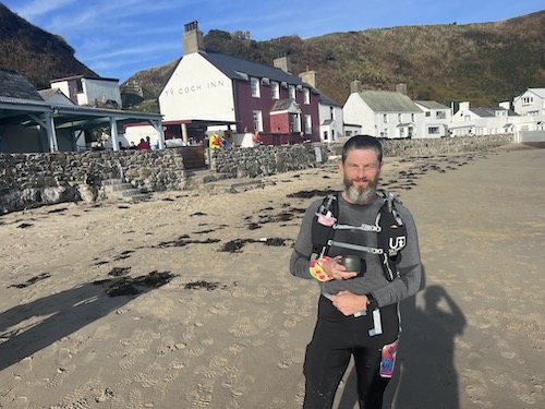 Running into Trouble: Garry standing on a sandy beach with houses behind him, holding a cup
