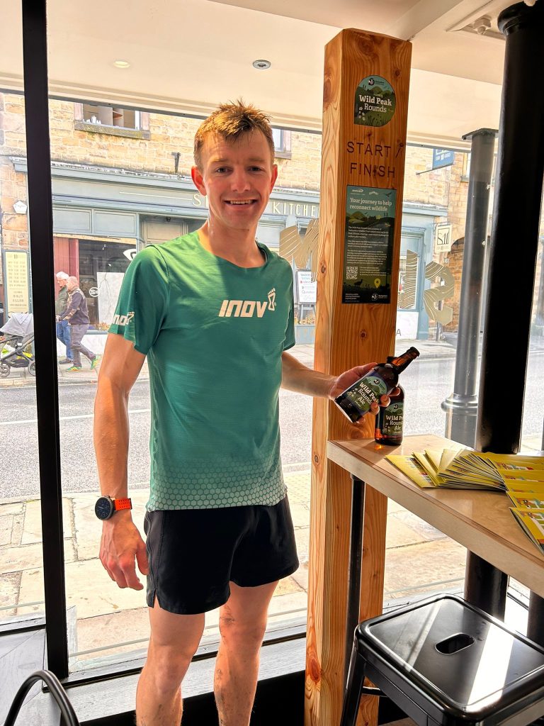 Wild Peak Round 120km Ultra Report - Jack Scott stands in a green inov8 t shirt in front of a large wooden post depicting the Wild Peak Rounds after finishing the 55km route