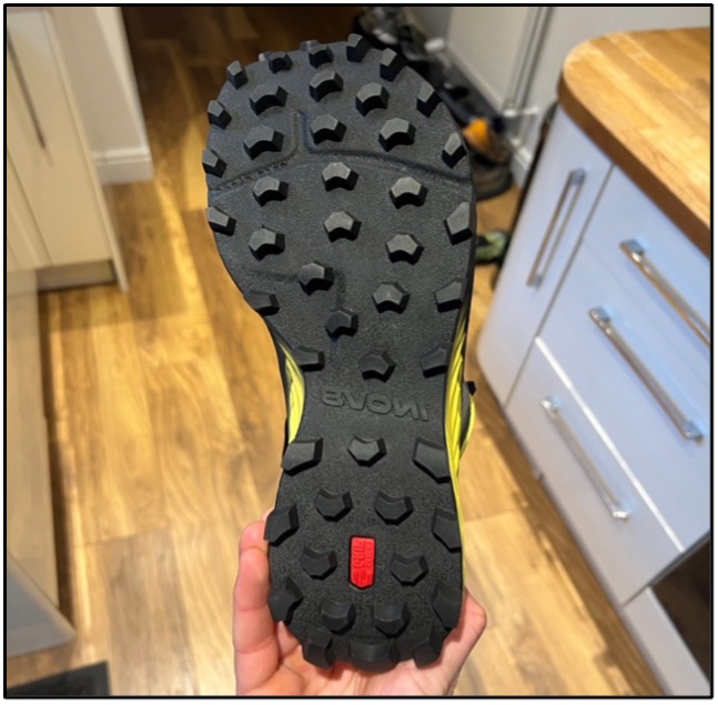A single inov8 MUDTALON SPEED being held upside down by a hand over a wooden floor