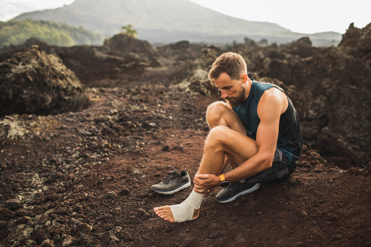 Male runner bandaging injured ankle. Injury leg while running outdoors. First aid for sprained ligament or tendon. Volcanic mountain view on background. Returning from injury in ultra running