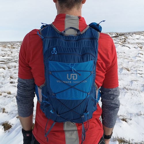 Ultimate Direction Adventure Vest Vesta 6.0 RunUltra Review A man wearing a blue vest over a red waterproof jacket, standing looking away from the camera on a snowy hillside