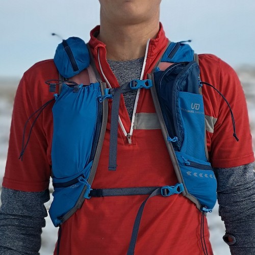 Ultimate Direction Adventure Vest Vesta 6.0 RunUltra Review A man wearing a blue vest over a red waterproof jacket, standing looking at the camera from the camera on a snowy hillside