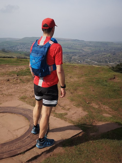 Ultimate Direction Adventure Vest/Vesta 6.0 RunUltra Review A man wearing a blue vest over a red t shirt, standing looking away from the camera on a hillside with a town in the distance