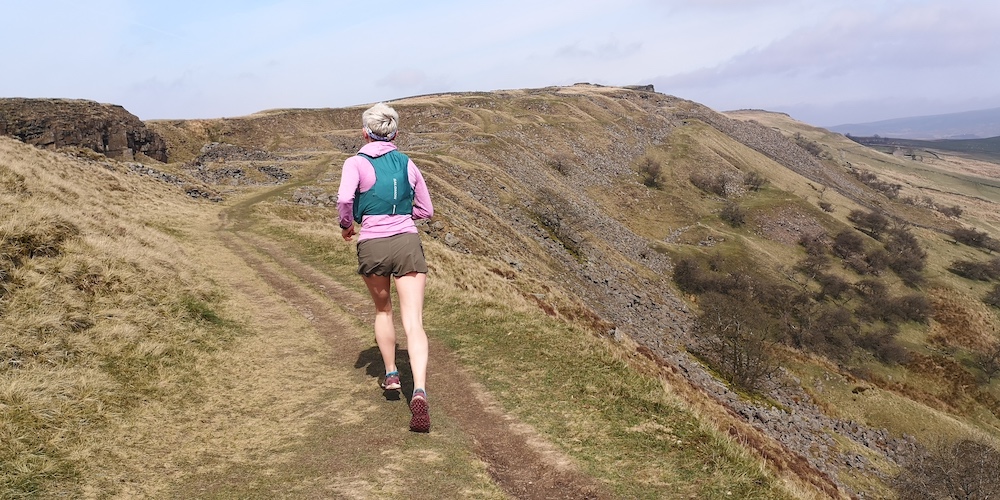Salomon Sense Aero 3" Shorts Review a woman running along a hillside with a valley beyond her wearing brown shorts, a pink top and a green vest.