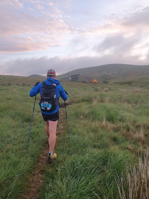 The Inaugural Summer Spine Challenger North A man walking with poles towards a hut with tents around it in a hilly landscape